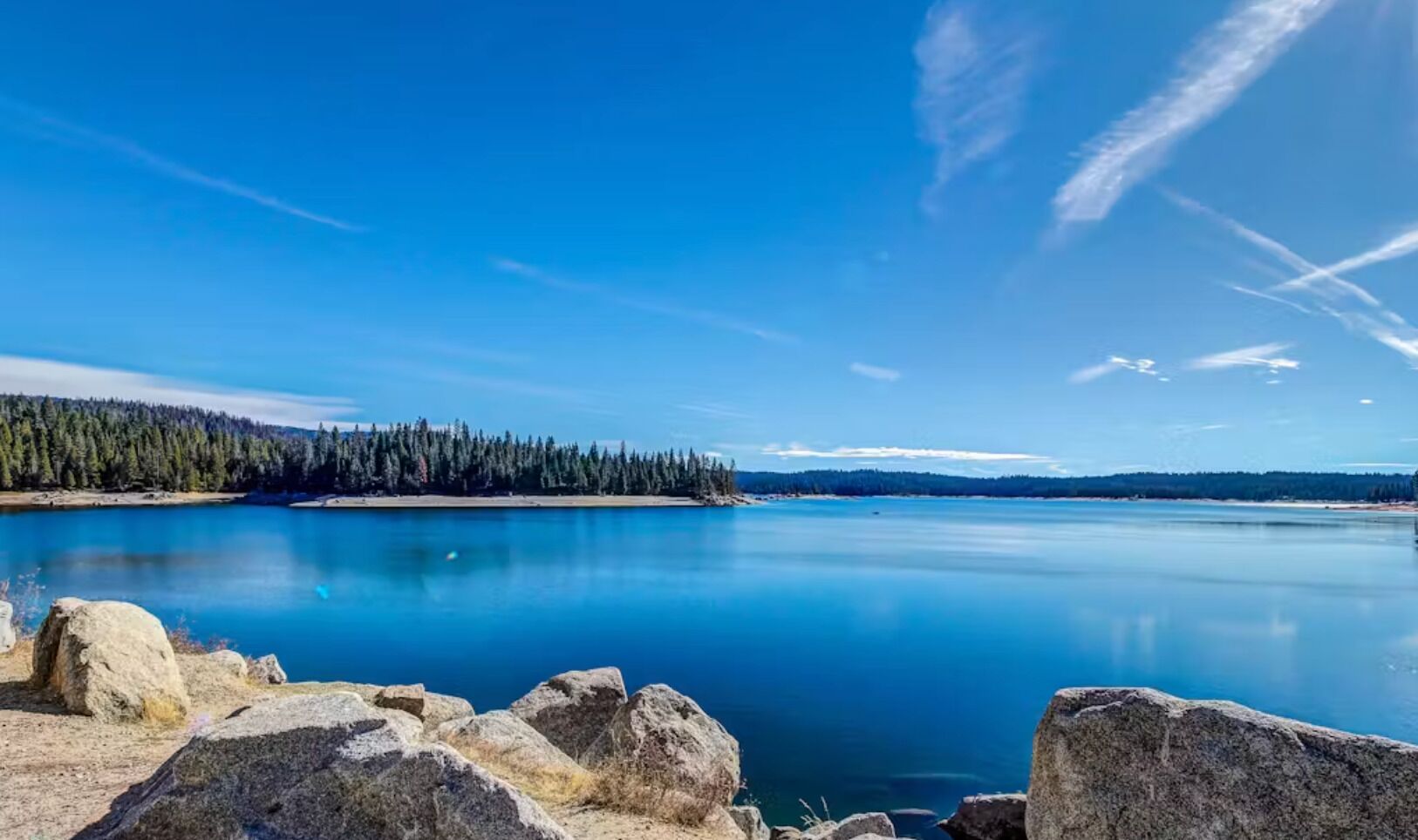 Un lago tranquilo con una orilla boscosa y grandes rocas al frente.