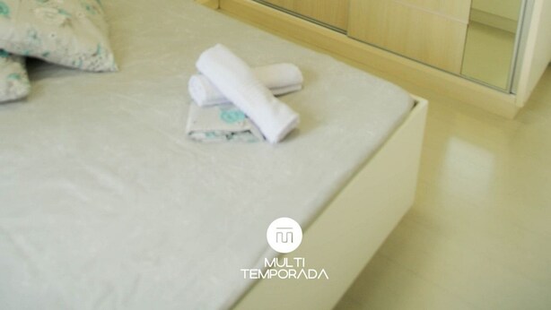 A modern bedroom with a bed, a bedside table, a mirror, and a sliding glass door leading to a bathroom. - Apartamento Centro de Imbituba - SC (Imbituba)