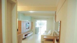 A modern living room with a flat-screen TV, a sofa, and a glass door leading to a balcony. - Apartamento Centro de Imbituba - SC (Imbituba)