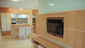 A modern kitchen with built-in appliances, a dining area with white chairs, and a large flat-screen TV mounted on a wooden wall. - Apartamento Centro de Imbituba - SC (Imbituba)