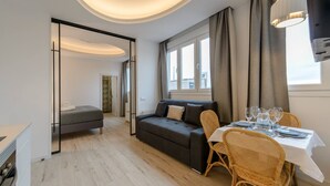 A modern living room with a sofa, dining table, and chairs. There is a large window with curtains, a ceiling light, and a door leading to another room. - GRAN VIA capitals, LiSBOA (Madrid)
