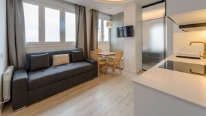 A modern living room with a sofa, a dining table with chairs, a flat-screen TV mounted on the wall, and a kitchen area with a refrigerator and a sink. - GRAN VIA capitals, LiSBOA (Madrid)