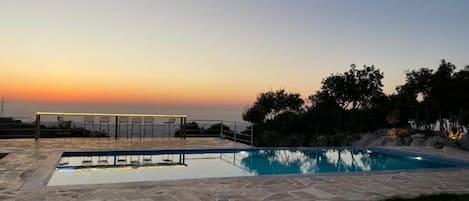 A rectangular swimming pool with a stone deck, surrounded by chairs and a railing, set against a sunset backdrop.