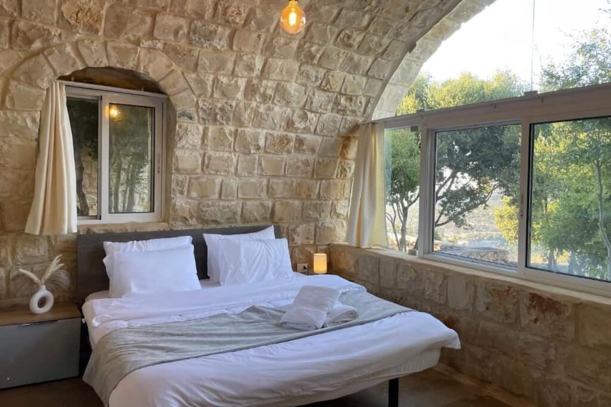 Un dormitorio con pared de piedra, una cama con ropa de cama blanca, una bombilla colgante, una ventana con vistas a árboles y una mesita de noche con lámpara.