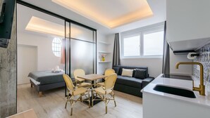 Ett modernt och kompakt bostadsutrymme med en soffa, ett matbord och stolar, ett litet kök samt ett stort fönster som släpper in naturligt ljus.