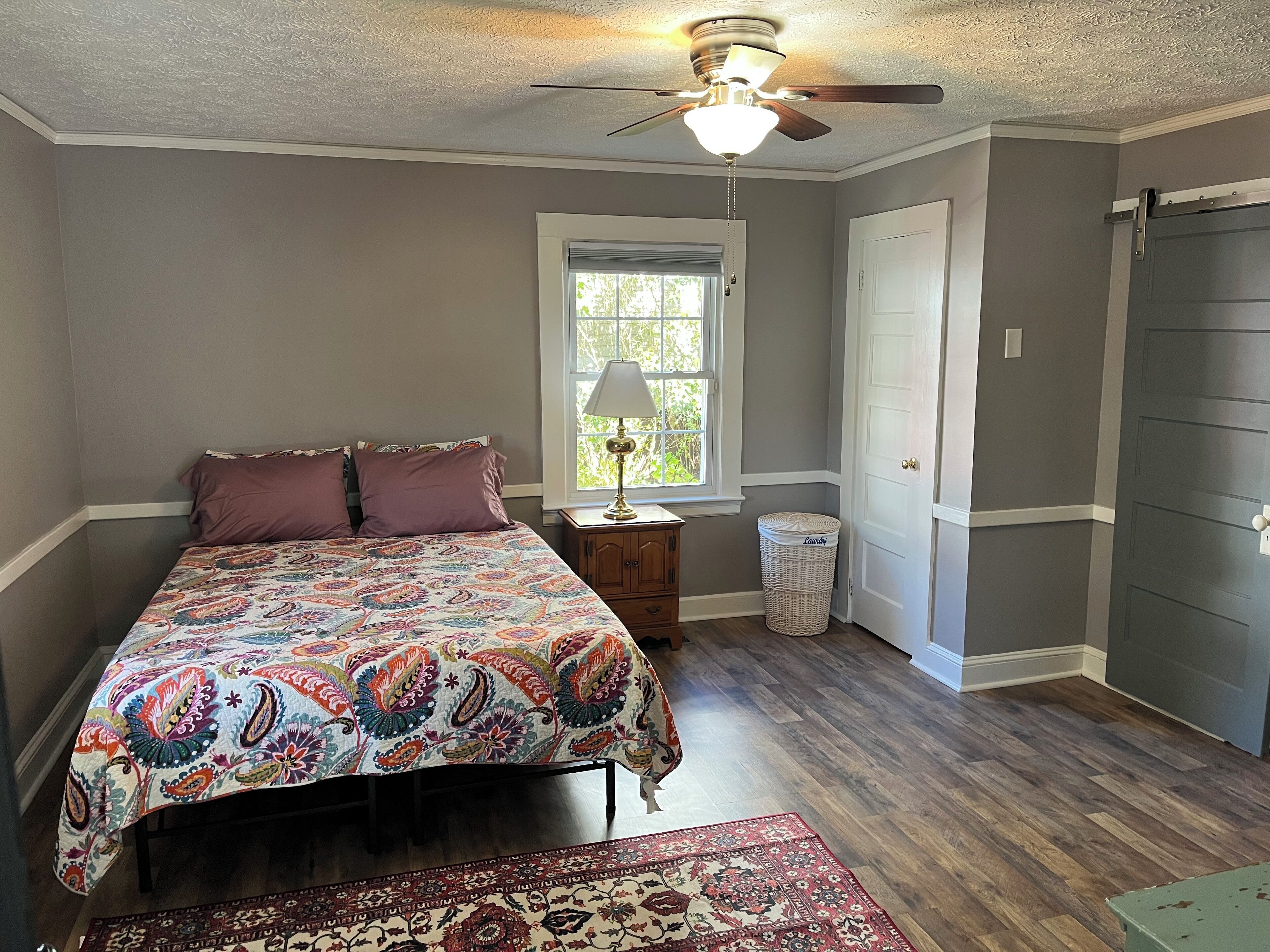 A bedroom with a bed, a nightstand, a lamp, a window, a door, and a ceiling fan.