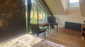 A room with a sloped ceiling, large windows, a bed, two chairs, a table, and a radiator. - Kick back and relax in this calm, stylish with views of beautiful Strathnairn. (Inverness)