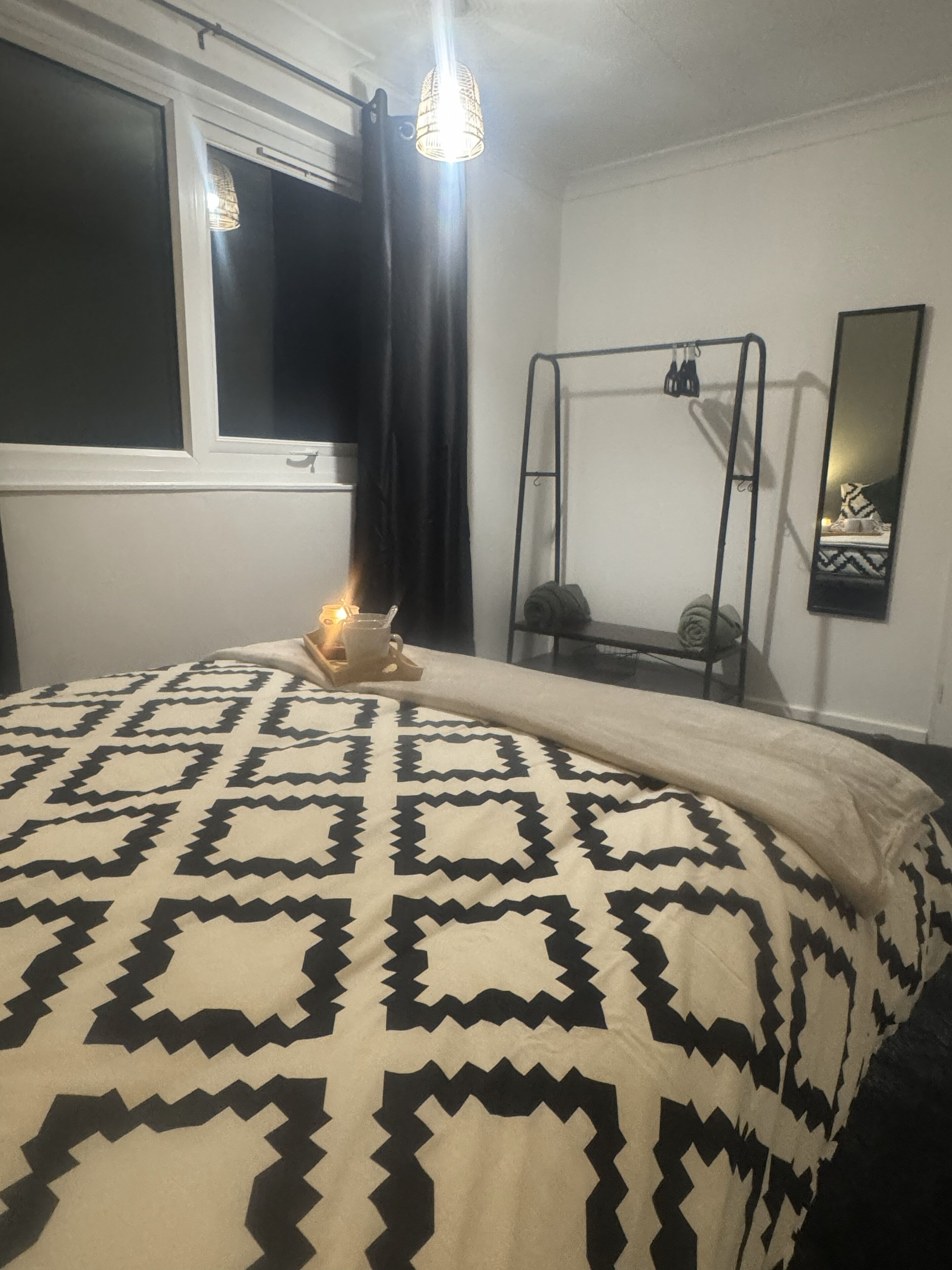 A bedroom with a bed, a window with curtains, a hanging light fixture, a metal rack, and a mirror on the wall.