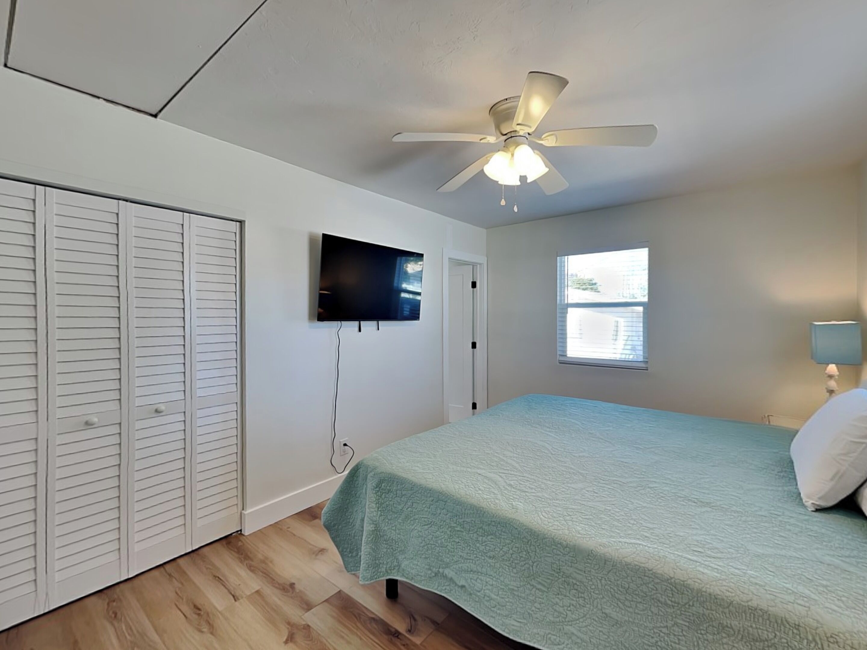 A bedroom with a ceiling fan, a bed with a green bedspread, a TV mounted on the wall, and a window with blinds.