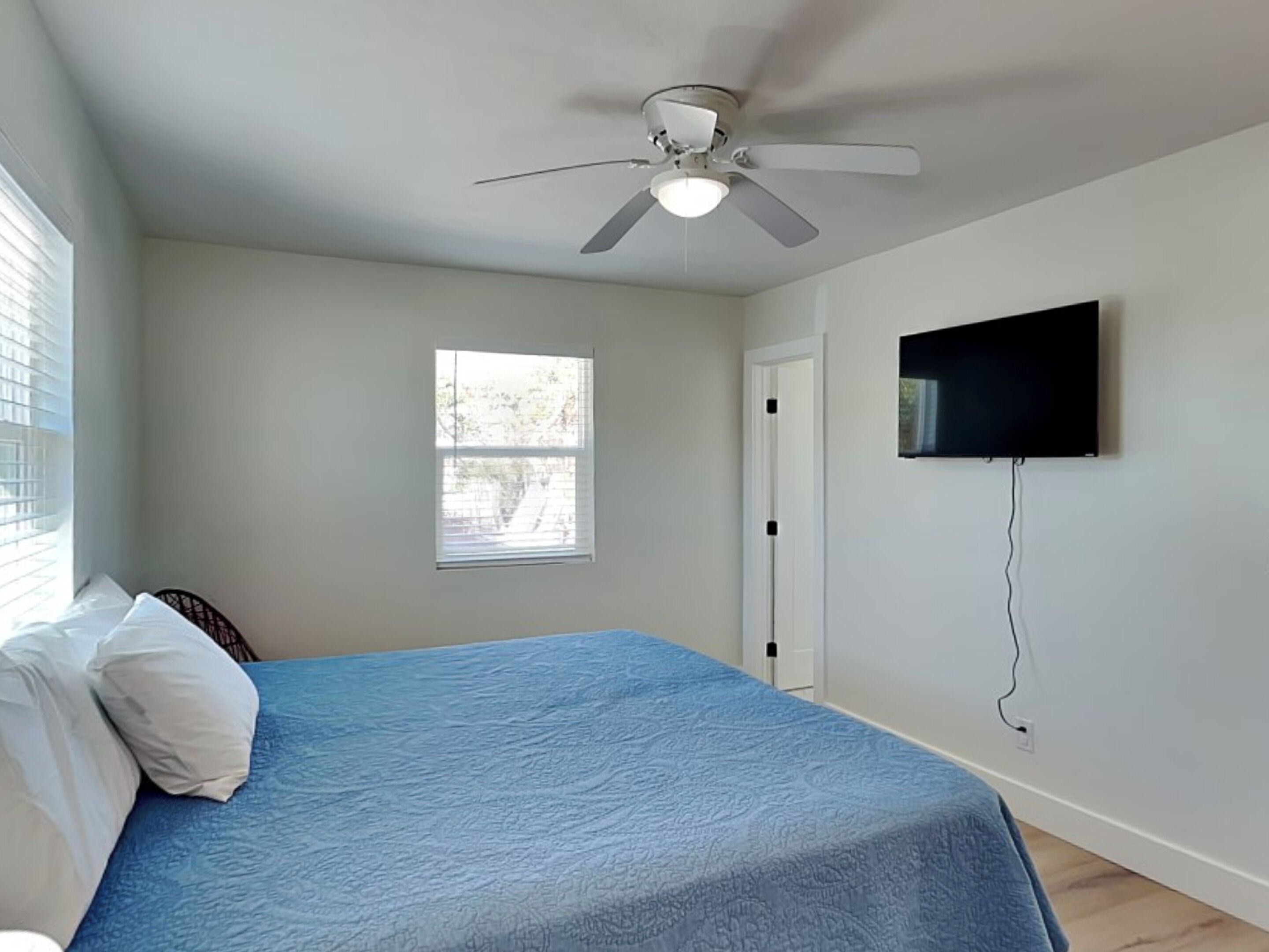 Ein Schlafzimmer mit Bett, Deckenventilator, Fenster, Tür und einem an der Wand befestigten Fernseher.