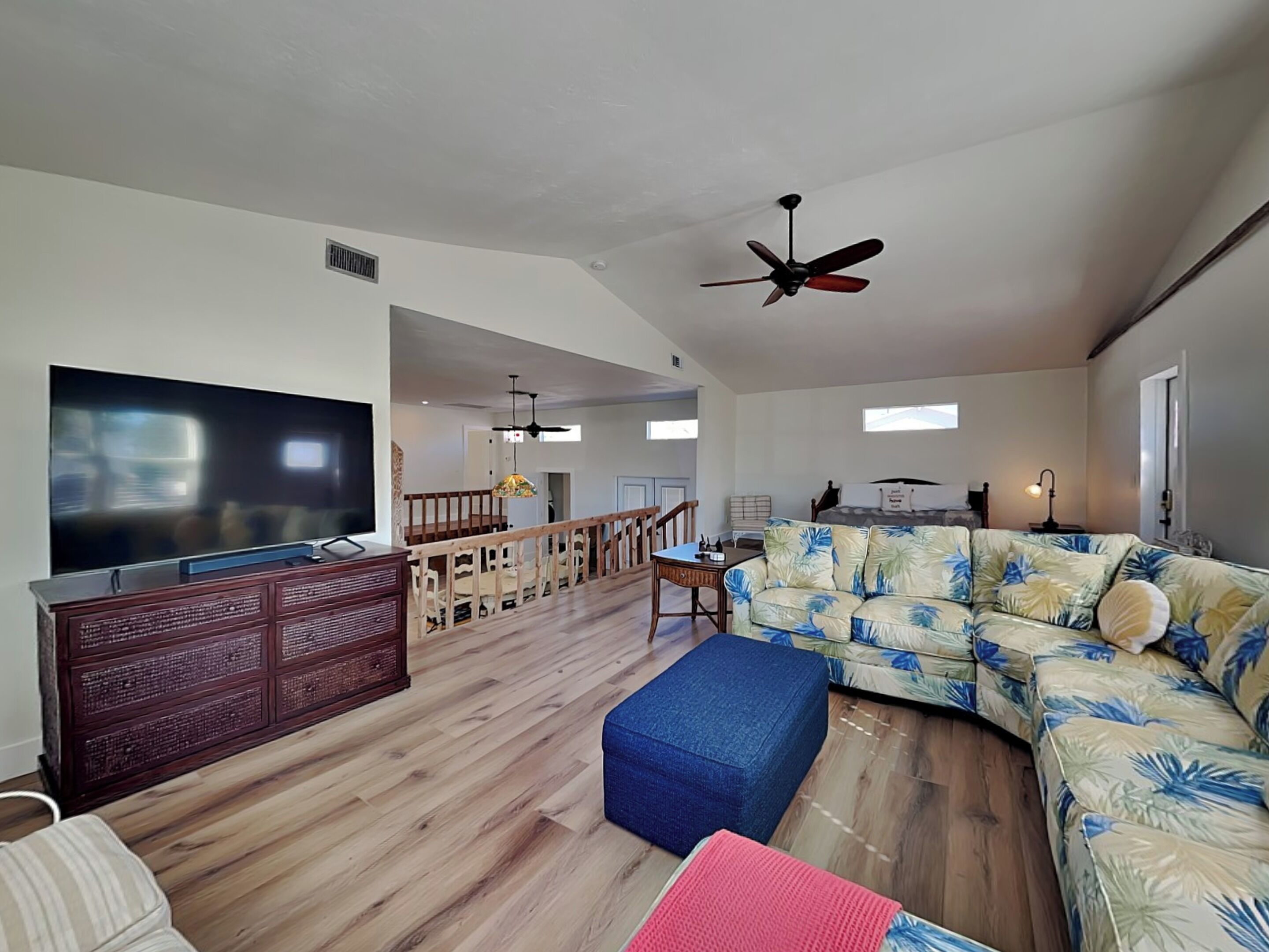 Ein geräumiges Wohnzimmer mit einem großen Flachbildfernseher, Holzböden, einem blauen Ottomanen, einem blumigen Sofa und einem Deckenventilator.