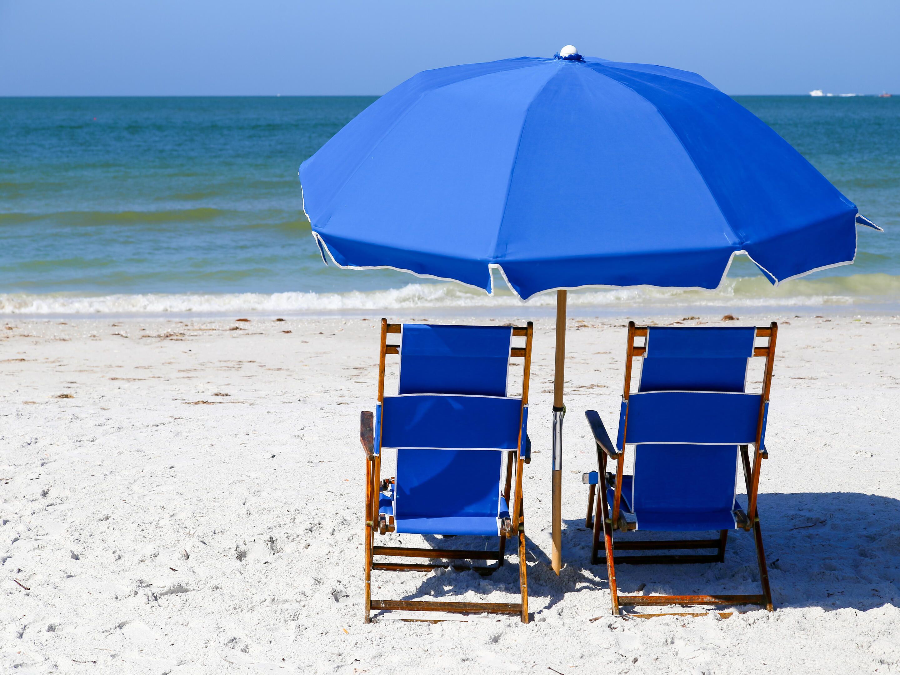 Two blue beach chairs and a blue umbrella on a sandy beach with the ocean in the background.