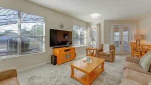 A living room with a flat-screen TV, wooden furniture, and large windows with blinds. - Poolside Living: Family Retreat 12 Mi to Disney (Davenport)
