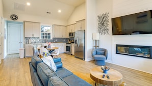A modern kitchen with white cabinets, a stainless steel refrigerator, and a microwave. There is a dining area with a table and chairs, and a living room with a blue sofa and a fireplace. - Lake Greenwood Access: Modern Escape w/ Patio! (Greenwood)