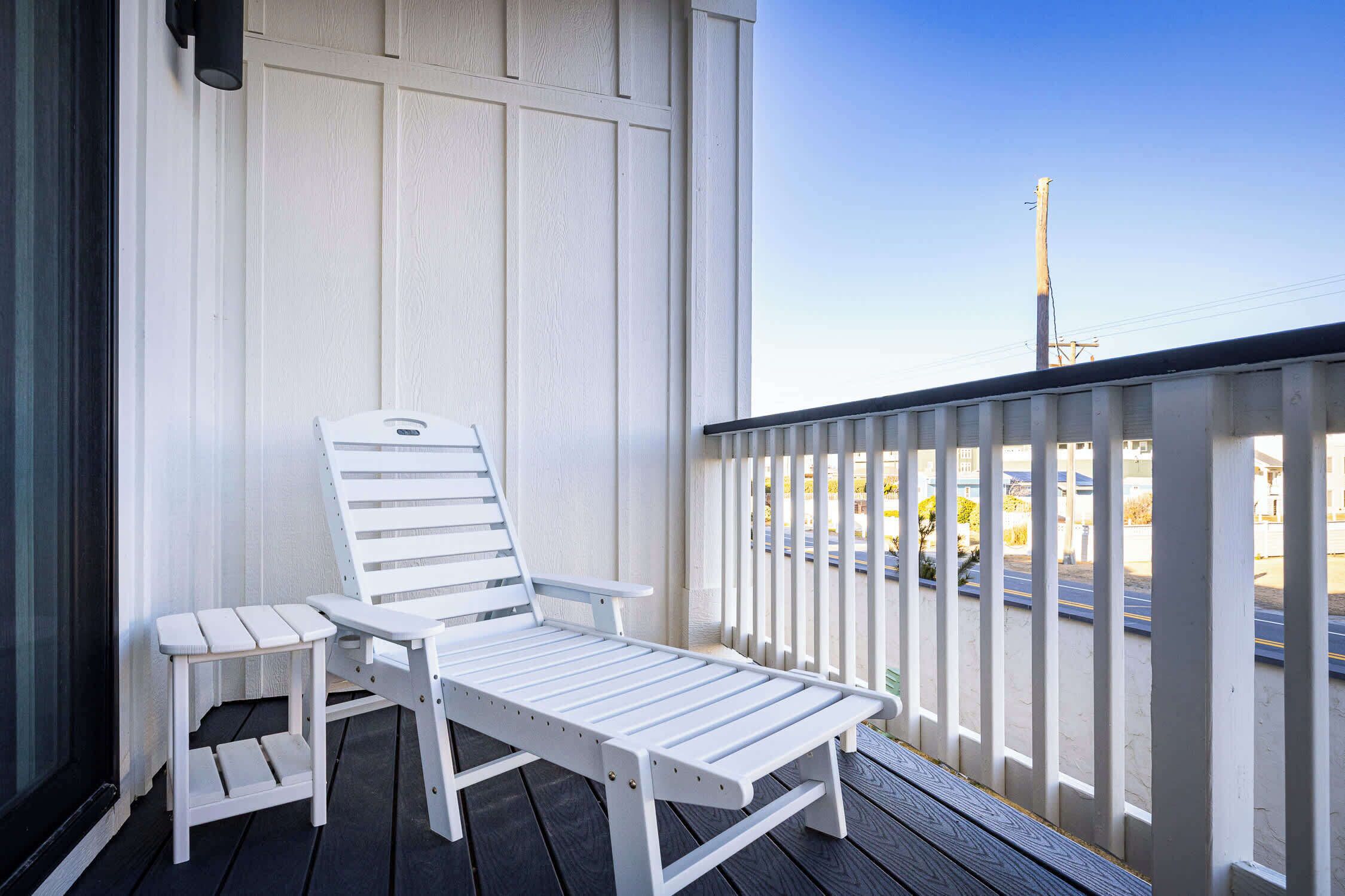A white outdoor seating area with a bench and a small table, a white railing, and a view of a road and buildings in the background.
