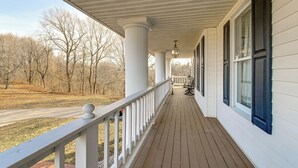A covered porch with white columns and a railing, overlooking a road and trees.
