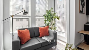 Moderni olohuone, jossa on harmaa sohva, oranssit koristetyynyt, lattiavalaisin ja suuri ikkuna, jossa on sälekaihtimet.