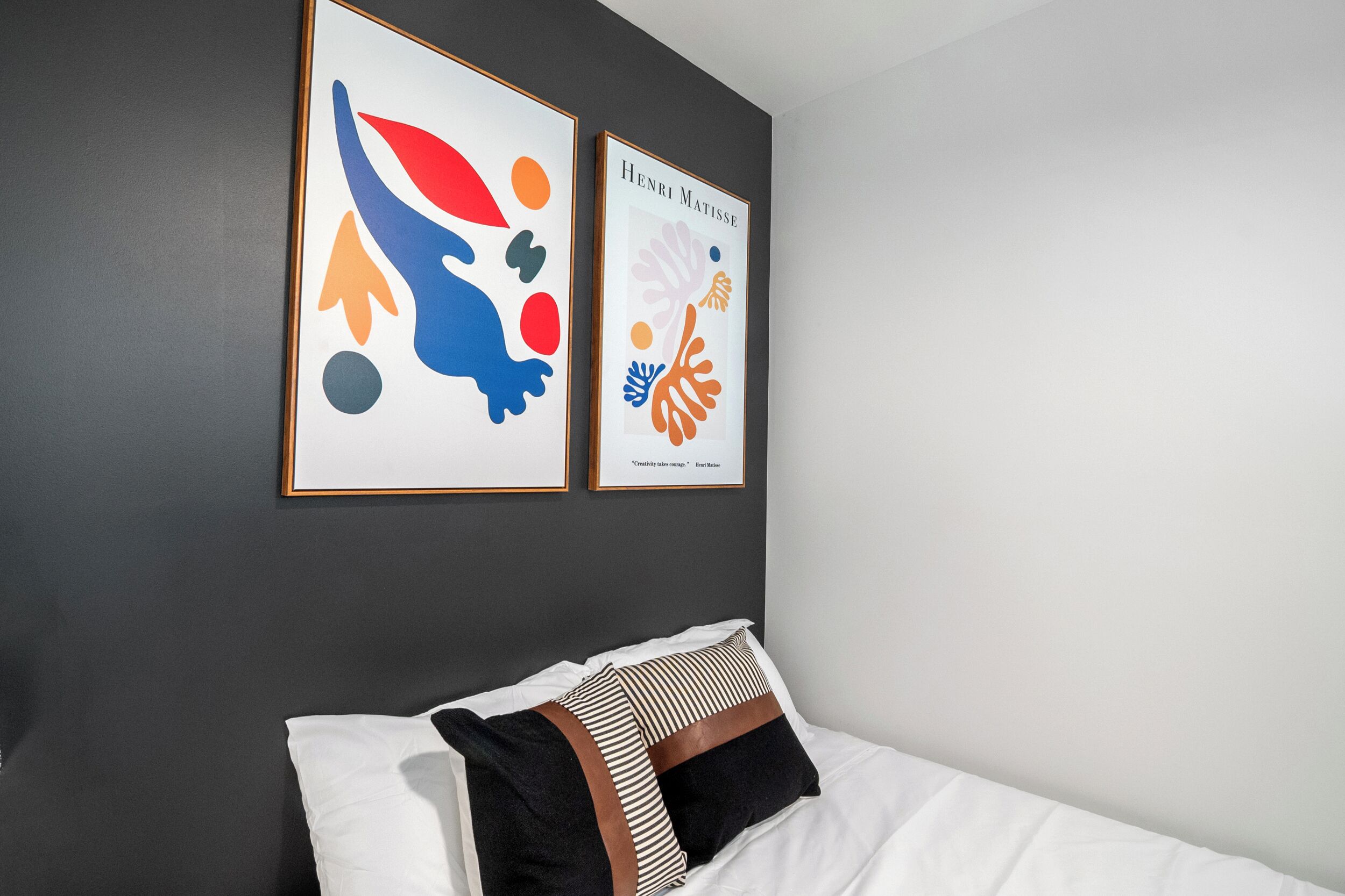 A neatly made bed with white and striped pillows, a dark wall, and two framed abstract artworks above it.