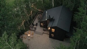 A cabin with a deck, surrounded by trees, and a fire pit on the ground.