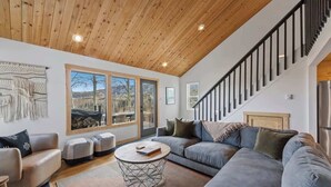 A modern living room with a wooden ceiling, large windows, a grey sofa, a round coffee table, and a staircase.