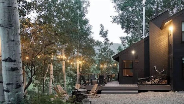 A modern cabin with a deck, surrounded by trees and string lights.