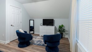 Une pièce avec une porte blanche, un ventilateur de plafond, un téléviseur à écran plat posé sur un meuble blanc, deux chaises bleues, un tapis bleu et une plante en pot.