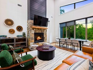 A modern living room with a stone fireplace, green upholstered chairs, a round coffee table, and a large window overlooking a forest. - Pinewood Haven | Swings, Playset, Arcade | Paved (Broken Bow)