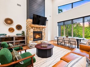 A modern living room with a stone fireplace, green upholstered chairs, a round coffee table, and a large window offering a view of the outdoors. - Pine Retreat | Hot Tub | Fire Pit Swings & Arcade (Broken Bow)
