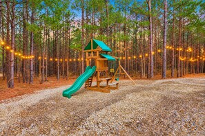 A wooden play structure with a slide and swing set in a forested area with gravel ground. - Pine Retreat | Hot Tub | Fire Pit Swings & Arcade (Broken Bow)