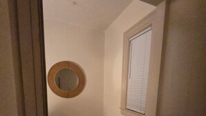 Un pasillo estrecho con suelo de madera, un espejo redondo pequeño en la pared y una ventana con persianas.