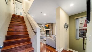 Escada de madeira que leva a uma sala de estar com lareira, um sofá e uma televisão na parede.