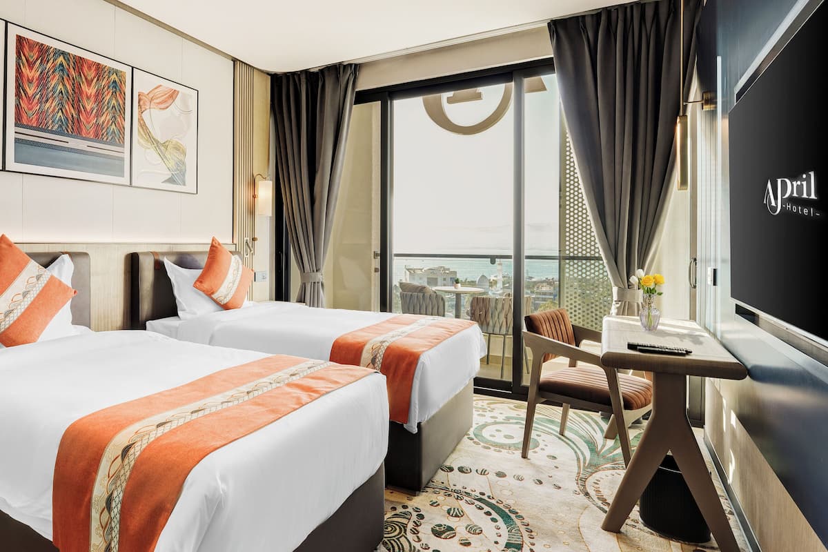 A hotel room with two beds, a desk with a chair, a TV, and a balcony with a view.
