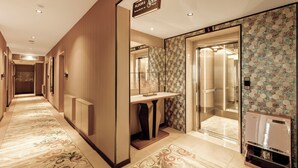 Un couloir d’hôtel avec un sol recouvert de moquette, un comptoir de réception, un ascenseur et un mur orné d’un papier peint à motifs floraux.
