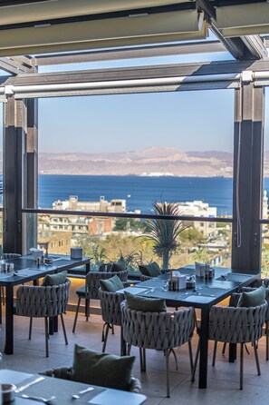 An outdoor dining area with tables and chairs, overlooking a coastal view with buildings and mountains in the distance. - April Hotel (Aqaba)