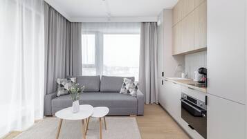 Ett modernt vardagsrum med en grå soffa, ett vitt soffbord och en köksdel med en spis och skåp.