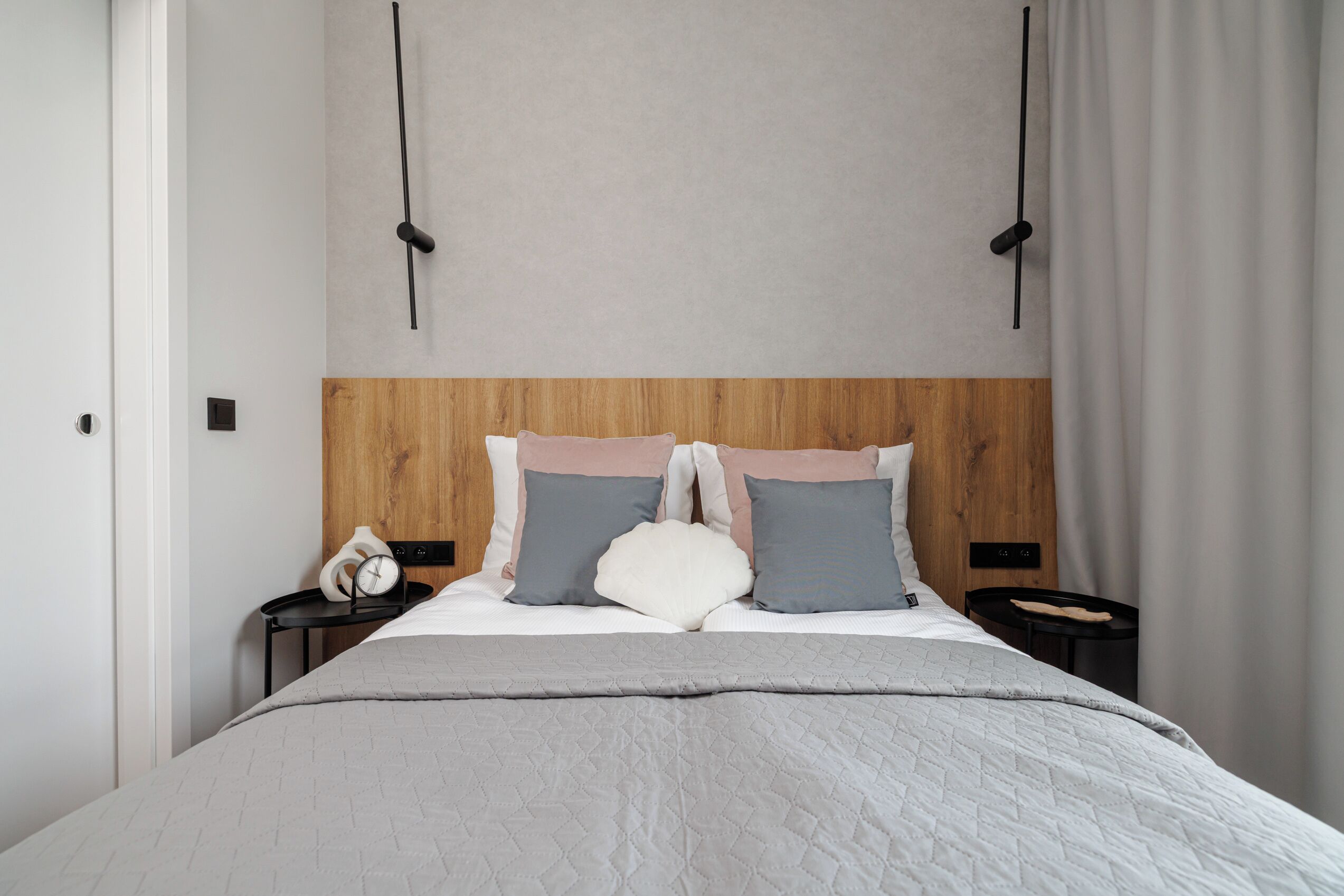 Un dormitorio moderno con cabecera de madera, cama con colcha gris, dos mesitas de noche con relojes y una cortina en la ventana.