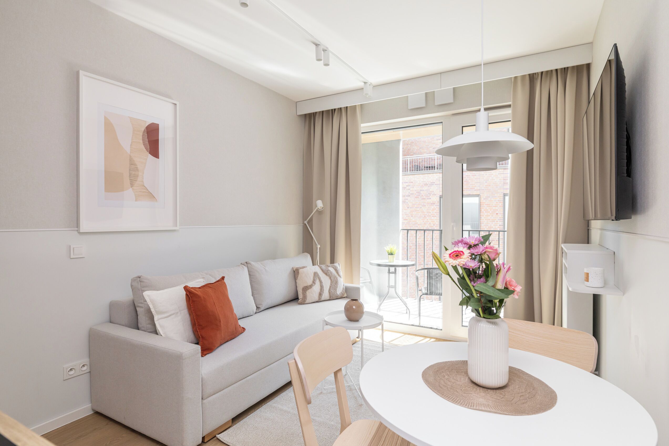 Ein modernes Wohnzimmer mit einer Couch, einem Esstisch mit Blumenvase, einem an der Wand montierten Fernseher und einem Balkon mit Blick auf ein Backsteingebäude.