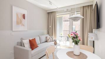 Ein modernes Wohnzimmer mit einer Couch, einem Esstisch mit Blumenvase, einem an der Wand montierten Fernseher und einem Balkon mit Blick auf ein Backsteingebäude.
