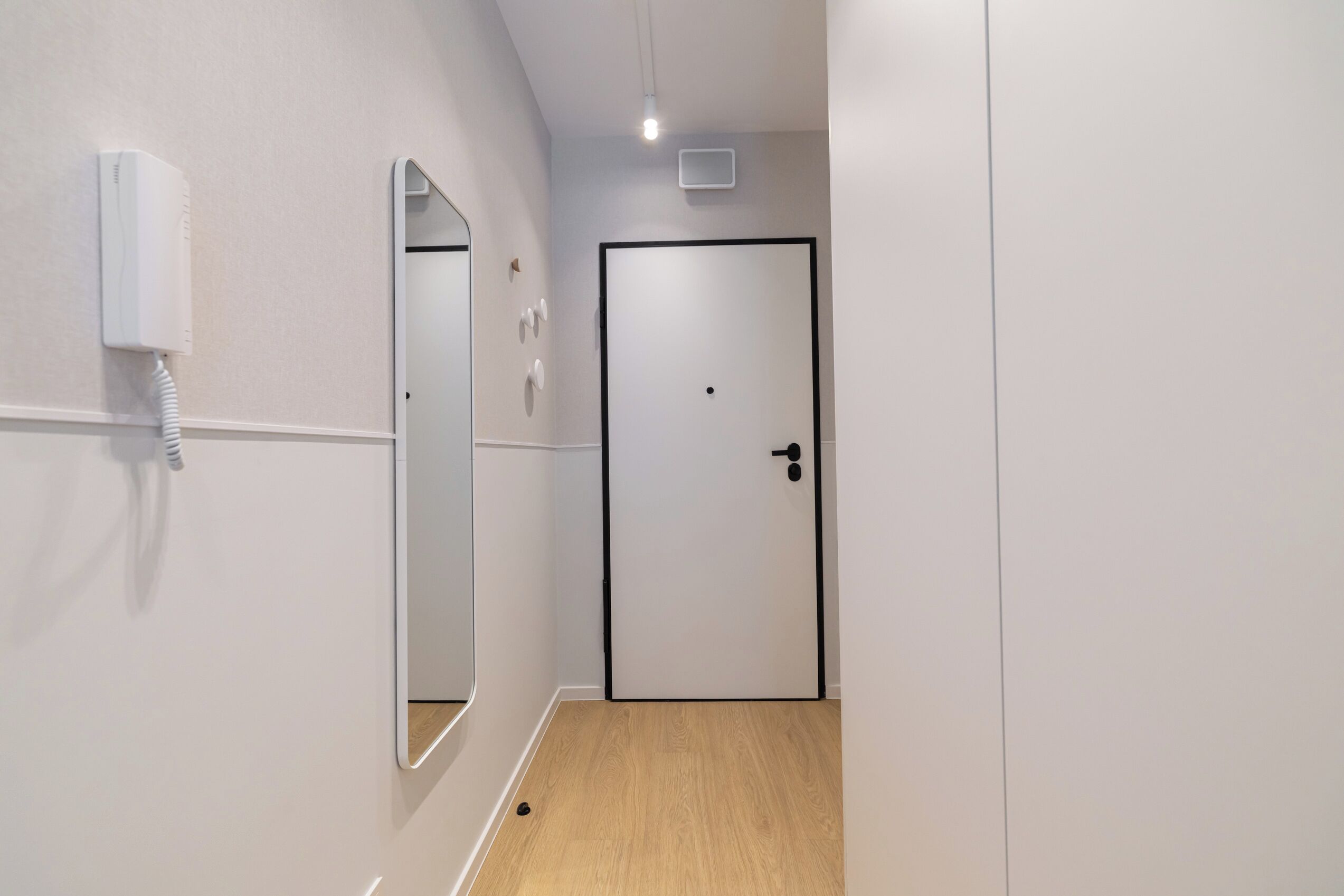 A hallway with a white wall, a door with a black frame, a mirror, and a wall-mounted phone.