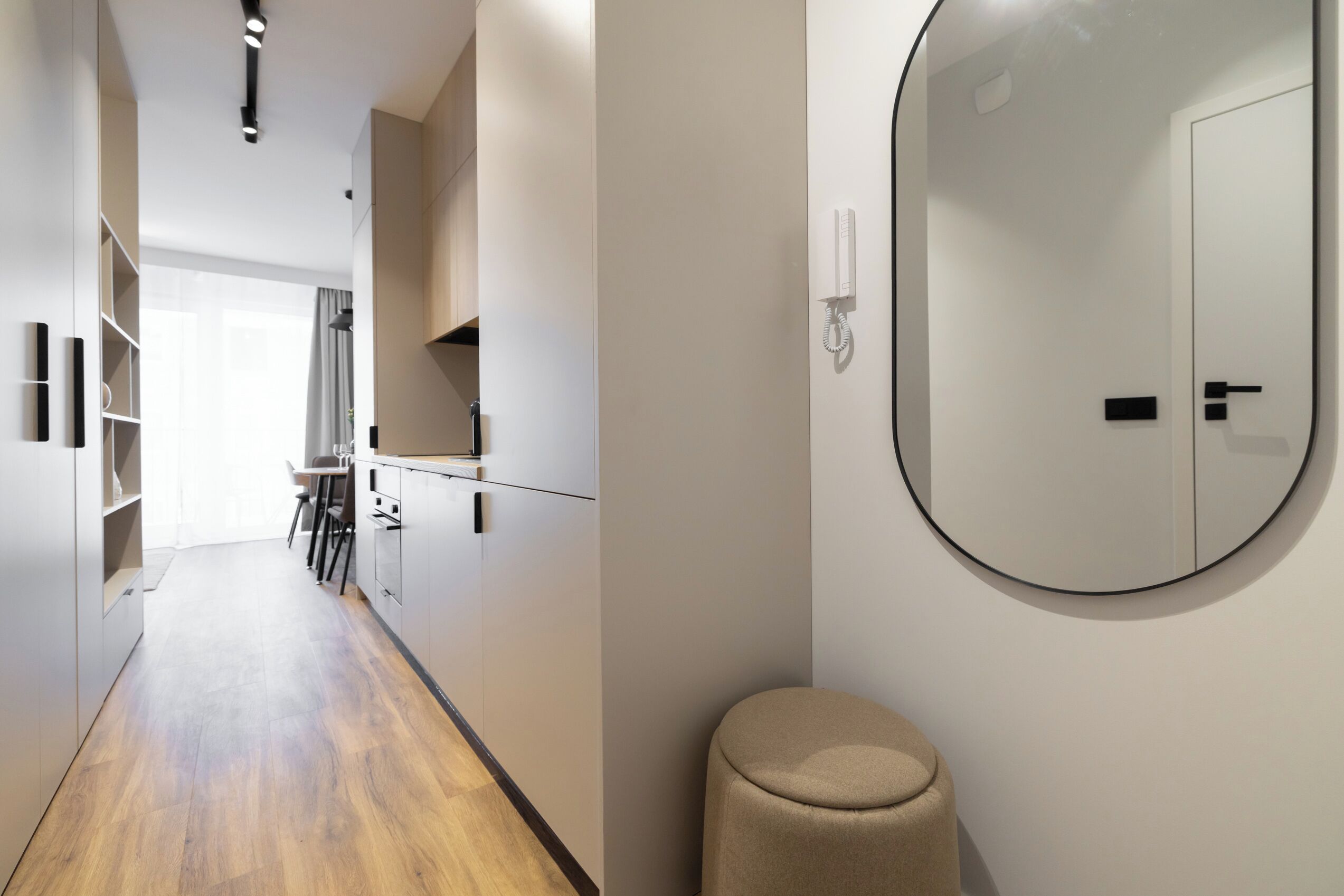 A modern hotel room with a wooden floor, a round mirror, a small stool, and a door with a handle.