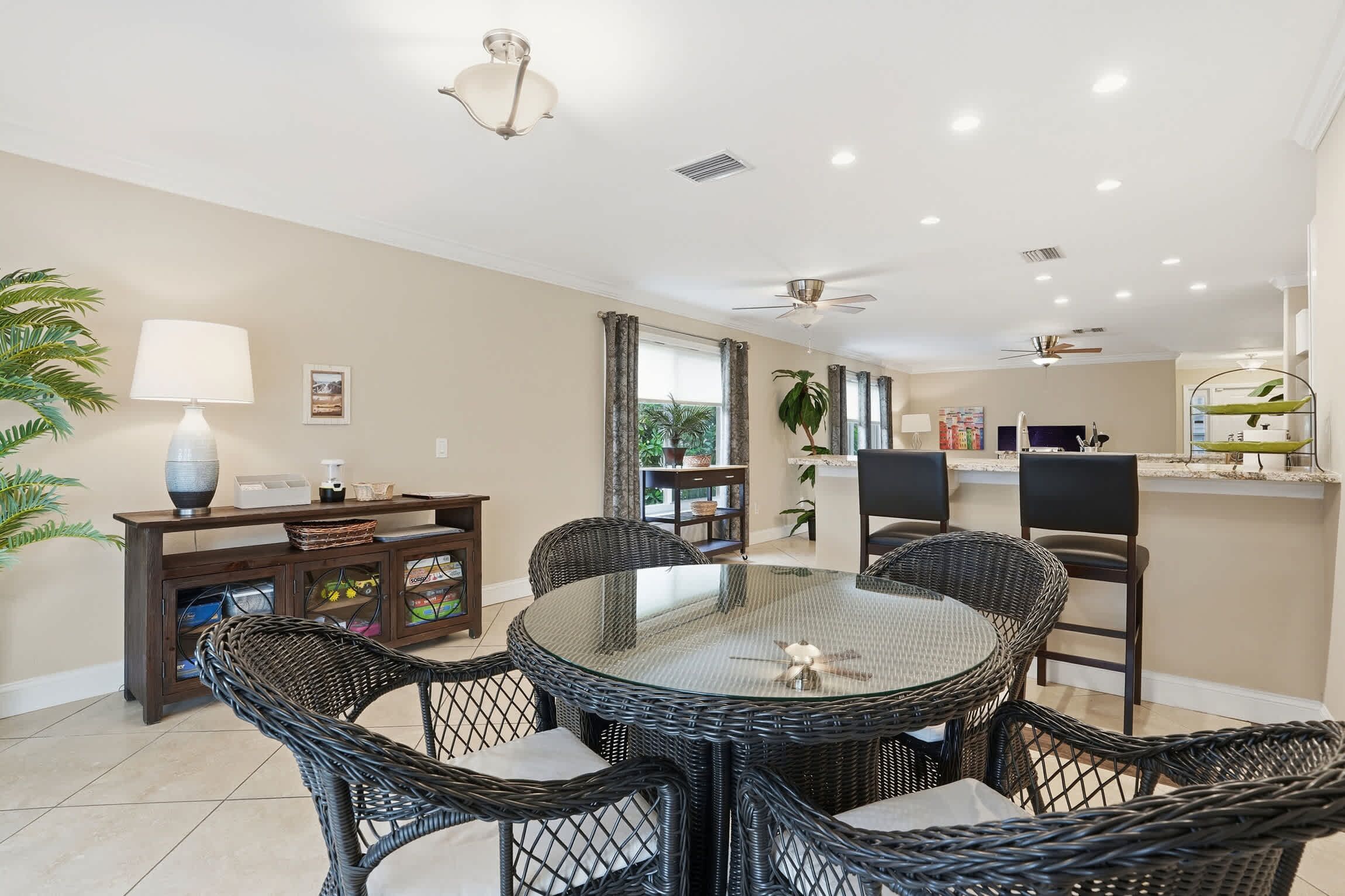 A dining area with a round glass table and wicker chairs, a dark wooden console table with a lamp, and a view of the kitchen area.