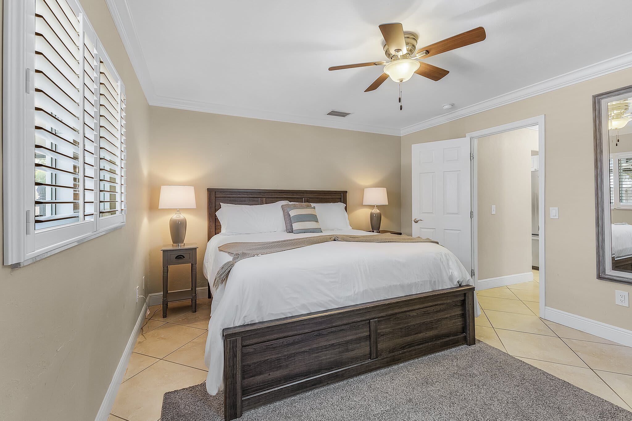 A bedroom with a bed, two bedside tables with lamps, a ceiling fan, and a large window with blinds.