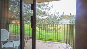 En balkong med ett metallräcke, en stol och utsikt över en gräsig gård med träd.