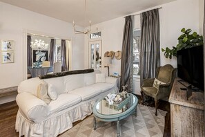 En stue med en hvid sofa, et glasbord, en lysekrone, et fjernsyn og en plante.