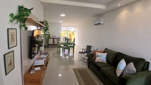Ein geräumiger Wohnbereich mit einem grünen Sofa, einem Holztisch und einem Esstisch mit Stühlen. Im Hintergrund ist eine Küche mit einem Fenster zu sehen, und links steht eine Pflanze.