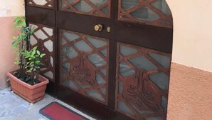 Une grande porte en métal, richement décorée de motifs complexes, avec un pot de fleurs placé à côté et un tapis rouge à l’entrée.