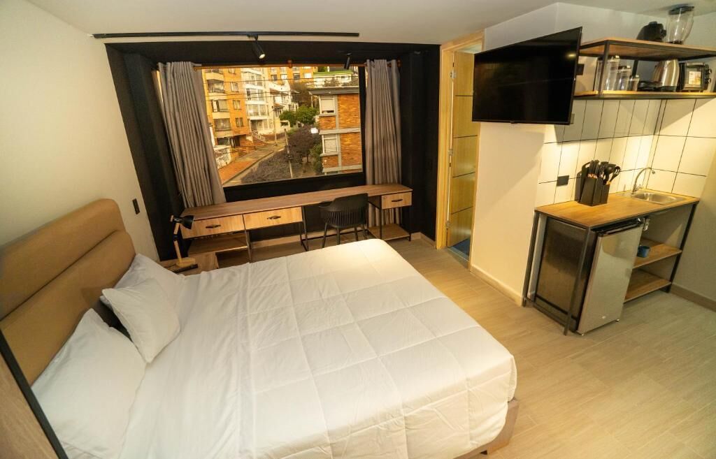 A bedroom with a bed, a desk, a chair, a TV, and a kitchen area with a sink and utensils.