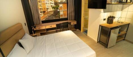 Un dormitorio con cama, escritorio, silla, televisión y una zona de cocina con pileta y utensilios.