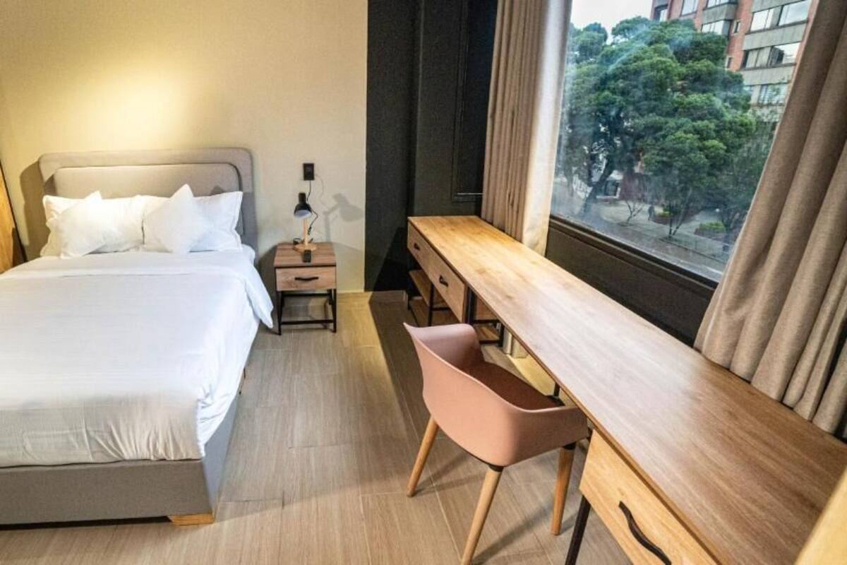 Hotelkamer met een bed, nachtkastje, lamp, bureau, stoel en een raam met uitzicht op gebouwen en bomen.