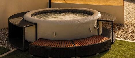 Ein Whirlpool mit Holzterrasse und Sonnenschirm, umgeben von einer Rasenfläche und einem Gebäude.